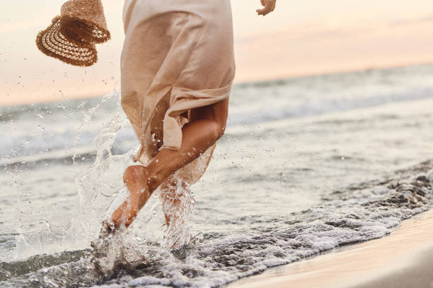 A woman runs along a beach in a flowing dress and straw hat, exemplifying carefree, body-positive fashion.