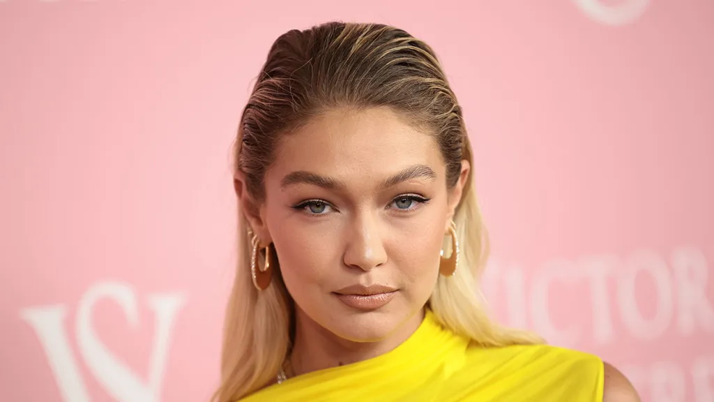 A fashion icon in a yellow top with slicked-back hair, embodying modern style on a pink background.